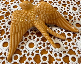 May include: A glossy, light brown ceramic wall ornament shaped like a bird in flight with outstretched wings. The bird features detailed, moulded feather textures across its body and wings, with a smooth, reflective finish throughout the piece.