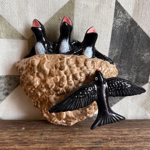 May include: A ceramic bird sculpture featuring a nest with three black birds with open pink beaks, and a single black bird in flight. The nest is a textured brown color, and the birds have white accents.