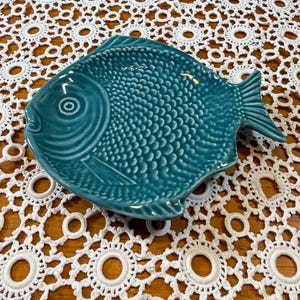Op de afbeelding: Een blauwgroene keramische schaal in de vorm van een vis met een gestructureerd schubbenpatroon over het lichaam, een duidelijk oog en een vin. Het oppervlak van de schaal is glanzend en glad, met een reliëfontwerp dat vissenschubben nabootst.