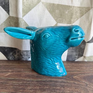 Pode incluir: Estatueta de cerâmica turquesa em forma de cabeça de burro. A cabeça do burro é detalhada com pelo texturizado e acabamento brilhante. A estatueta está colocada sobre uma superfície de madeira e um fundo de tecido estampado.