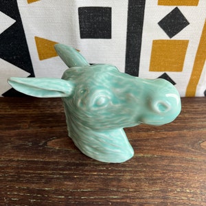 May include: A turquoise ceramic moose head ornament. The moose head is glossy and detailed, with visible ears, eyes, and a snout. The ornament is set against a backdrop of a patterned fabric with black, white, and gold geometric shapes, and a wooden surface.