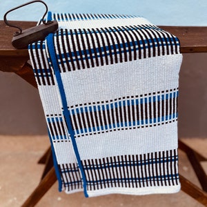 May include: A white, black, and blue striped woven throw blanket with a blue border. The blanket is folded and draped over a wooden ironing board.