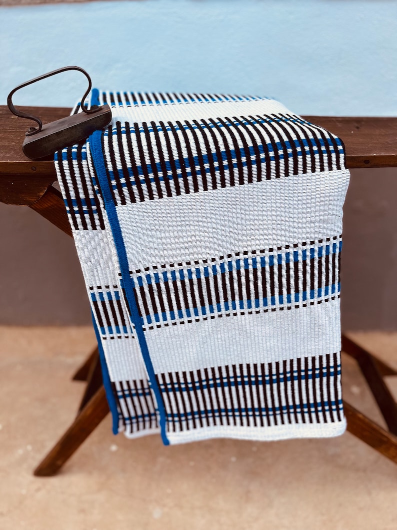 May include: A white, black, and blue striped woven throw blanket with a blue border. The blanket is folded and draped over a wooden ironing board.