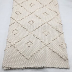 May include: A cream-colored woven rug with a diamond pattern. The rug has a textured surface with raised bumps.