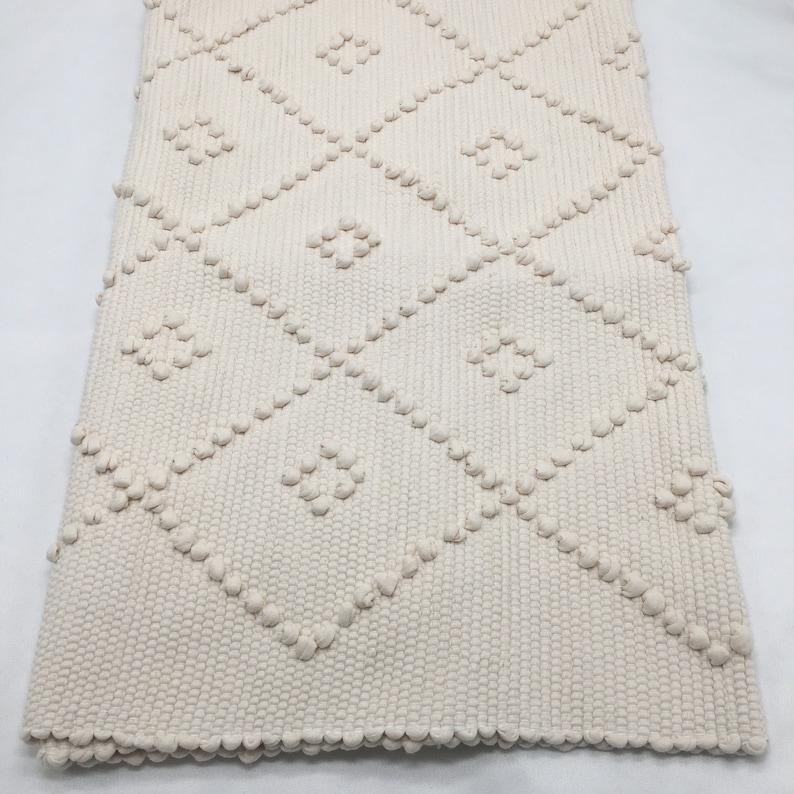 May include: A cream-colored woven rug with a diamond pattern. The rug has a textured surface with raised bumps.