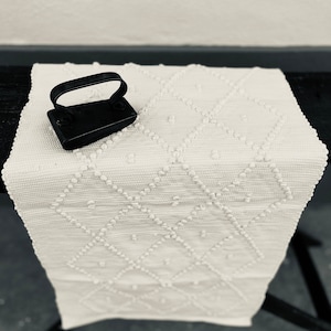 May include: A white crocheted rug with a diamond pattern and a small black iron. The rug has a raised texture and is perfect for adding a touch of rustic charm to any room.