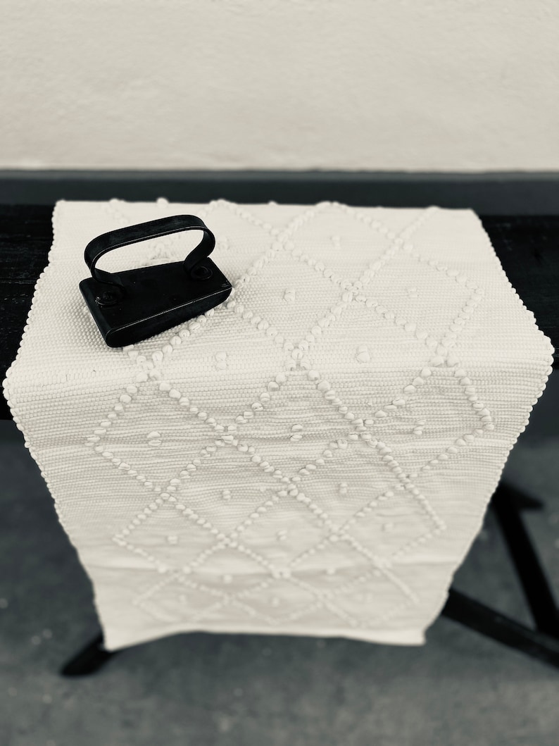 May include: A white crocheted rug with a diamond pattern and a small black iron. The rug has a raised texture and is perfect for adding a touch of rustic charm to any room.