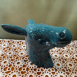 May include: A teal ceramic sculpture shaped like the head of a donkey, featuring a glossy finish, textured fur details, one prominent ear extending to the left, and a defined snout with a closed mouth, captured from a side profile angle.