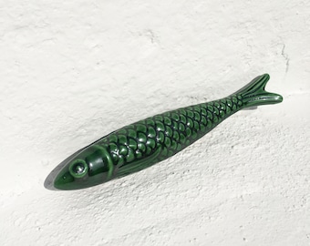 Puede incluir: Una figurita de pescado de cerámica verde con un patrón de escamas detallado. El pez mide aproximadamente 15 cm de largo y tiene una forma realista.