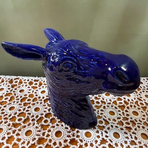 May include: A glossy, deep cobalt blue ceramic sculpture shaped like the head of a donkey. The piece features detailed molded textures representing the animal's fur, prominent ears, and facial features, finished with a smooth, reflective glaze.