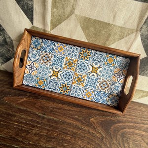 May include: Wooden serving tray with a rectangular shape and integrated handles. The tray's interior features a decorative tile design in blue, white, and yellow. The tray is set on a dark wooden surface.