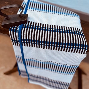 May include: White woven throw blanket with blue and black stripes. The blanket is folded and draped over a wooden surface.