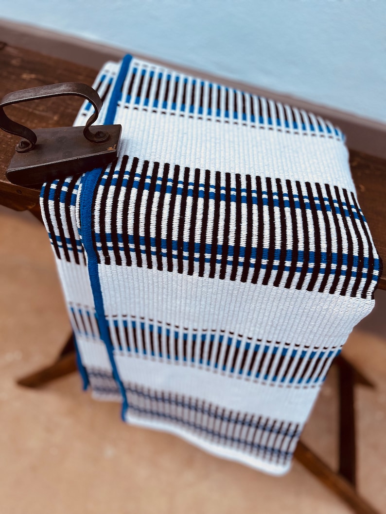 May include: White woven throw blanket with blue and black stripes. The blanket is folded and draped over a wooden surface.