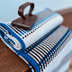 May include: A white and blue striped throw blanket with a black iron vintage clothes iron on top. The blanket has a blue border.