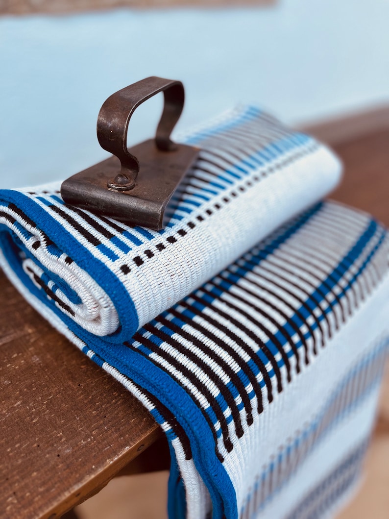 May include: A white and blue striped throw blanket with a black iron vintage clothes iron on top. The blanket has a blue border.