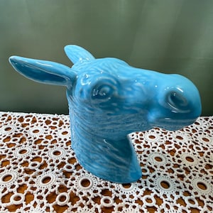 Pode incluir: Uma escultura de cerâmica azul brilhante em forma de cabeça de burro. A peça apresenta um acabamento brilhante e reflexivo com detalhes moldados que destacam as orelhas, os olhos e o focinho do animal, capturando uma textura realista.