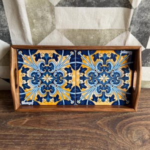 Puede incluir: Bandeja de madera con dos baldosas de cerámica decorativas. Las baldosas presentan un diseño floral azul, amarillo y blanco. La bandeja tiene forma rectangular con asas. La palabra "Portugal" está impresa en las baldosas.
