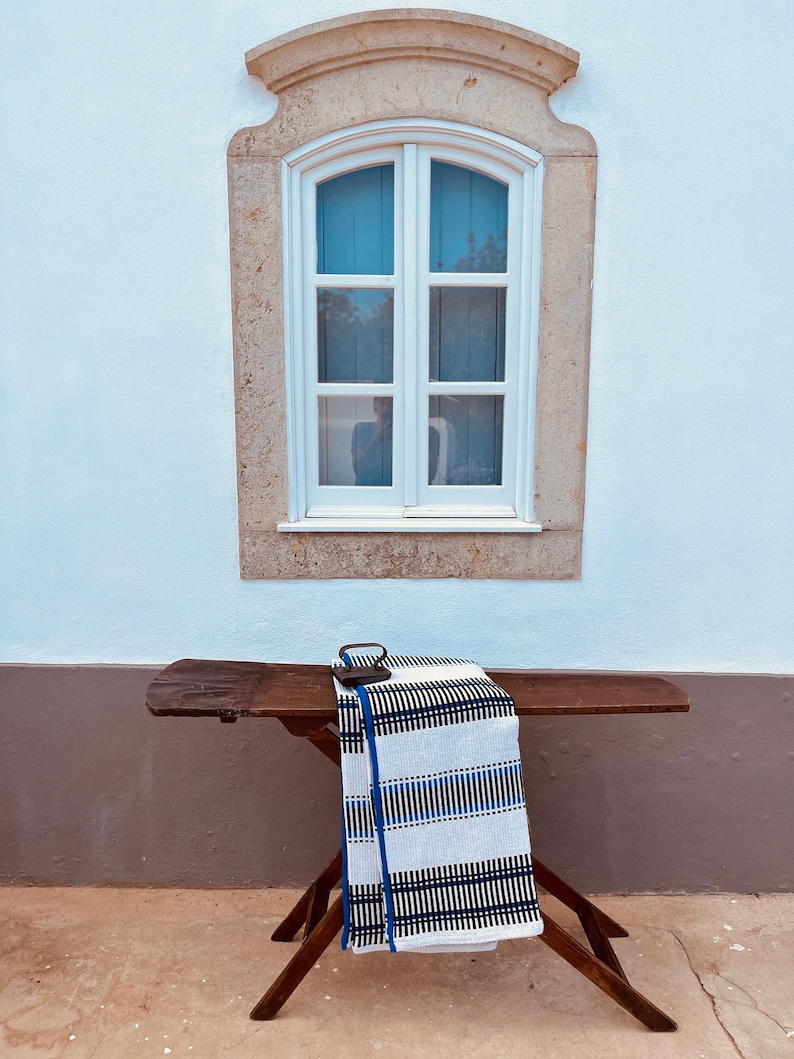 May include: A white wooden ironing board with a black and white striped blanket folded on top. The ironing board is set up in front of a white wall with a window.