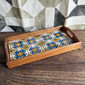 Puede incluir: Bandeja de madera rectangular con asas. La bandeja presenta un inserto de azulejos decorativos con un patrón floral azul, amarillo y blanco. La madera tiene un acabado cálido y natural. Mide unos 30 cm de largo.