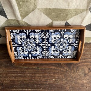 May include: Wooden serving tray with a rectangular shape and integrated handles. The tray features four square ceramic tiles with a blue and white floral pattern. The tray is set on a dark wooden surface.