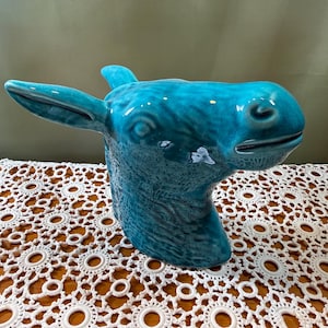 Pode incluir: Uma escultura de cerâmica turquesa em forma de cabeça de burro, com acabamento brilhante e um padrão de esmalte craquelado visível. A peça apresenta orelhas proeminentes, traços faciais definidos e um focinho liso e arredondado em azul.