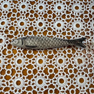 Könnte beinhalten: Eine Keramikfischfigur mit einem grau-weißen Schuppenmuster, einer dunkelgrauen Flosse und einem dunkelgrauen Schwanz. Der Fisch ist in einer horizontalen Schwimmposition dargestellt, mit einem detaillierten Auge und einer glatten, glänzenden Oberfläche.