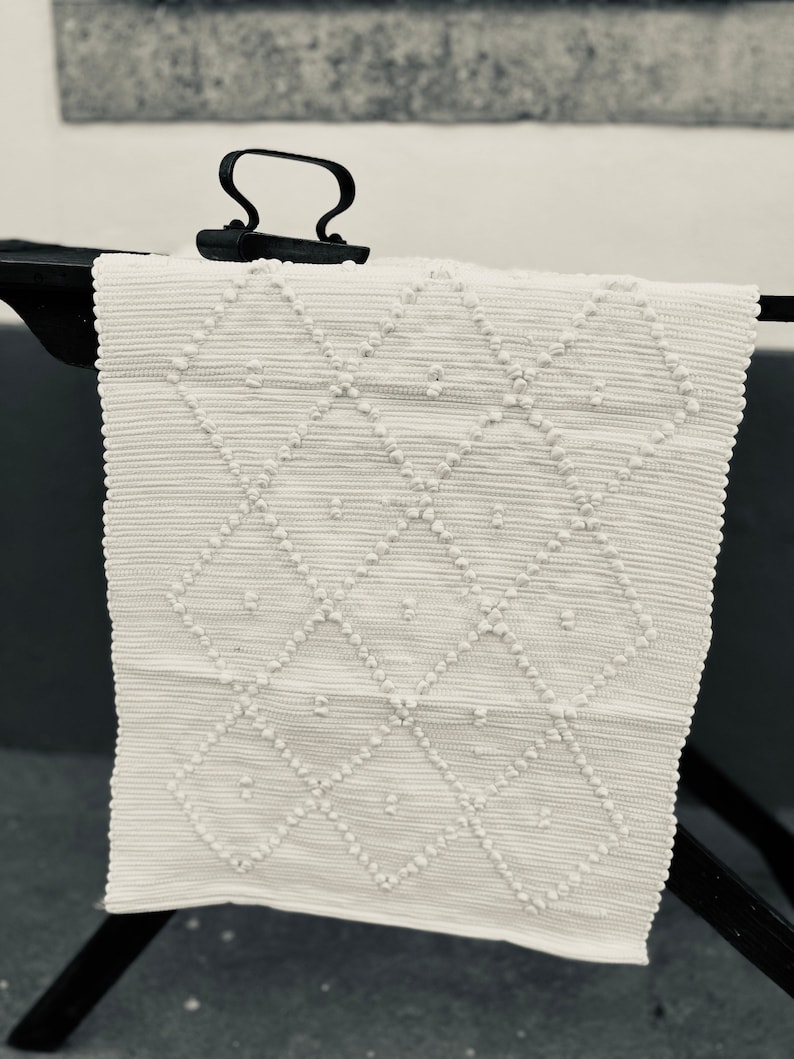 May include: A white woven rug with a diamond pattern and raised dots. The rug is hanging on a black metal rack.
