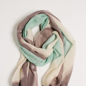 Puede incluir: Bufanda ligera en tonos marrón, verde menta y crema. La bufanda tiene forma rectangular con bordes deshilachados. Los colores están dispuestos en rayas y se mezclan, creando un efecto acuarela suave. Este accesorio es adecuado para cualquier estación.