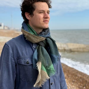 May include: A man wearing a blue denim jacket with a multi-colored scarf. The scarf has a geometric pattern in shades of green, blue, and brown. The man is standing on a beach with the ocean in the background.