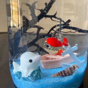May include: A clear glass jar with a blue sand base, a white dolphin figurine, a pink boat, a seashell, and a red and silver fish hanging from the top.