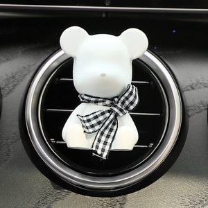 May include: White ceramic bear car air freshener with a black and white checked scarf. The bear is sitting in a car's air vent.