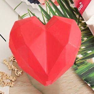 May include: A red geometric heart-shaped object made of a smooth, solid material. The heart is sitting on a wooden surface.