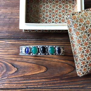 Può includere: Un braccialetto d'argento con cinque pietre preziose di forma ovale. Le pietre preziose sono verdi, nere e blu.