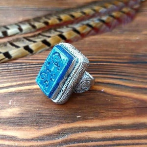 May include: A silver ring with a blue stone set in a rectangular shape. The stone has Arabic script engraved on it.