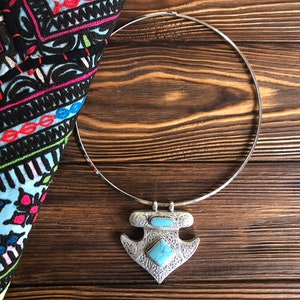 Puede incluir: Un collar choker plateado con un colgante de ancla con piedras turquesa. El collar está sobre una superficie de madera.