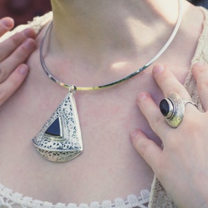 Puede incluir: Un collar choker plateado con un gran colgante triangular que presenta un diseño intrincado y una piedra negra. El colgante está unido al collar con una cadena plateada. El collar lo lleva una persona con dos anillos plateados en los dedos, uno con una piedra negra y el otro con una piedra negra.