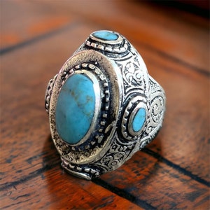 Può includere: Un anello d'argento con una grande pietra ovale turchese e due pietre turchesi più piccole. L'anello è finemente dettagliato con motivi floreali.