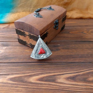 May include: A silver pendant with intricate designs and a red gemstone in the center. The pendant is sitting on a wooden surface next to a wooden box with a metal handle.