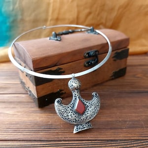 Puede incluir: Un collar plateado con un colgante de diseño ornamentado y una gema roja. El collar descansa sobre un cofre de madera con herrajes metálicos, sobre un fondo cálido y neutro.