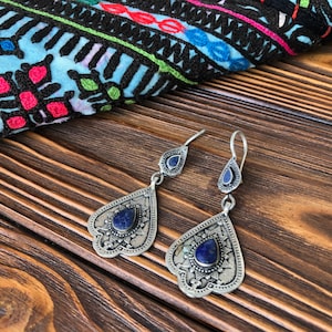 May include: Silver-toned drop earrings with a teardrop design. Each earring features a central, deep blue stone surrounded by an ornate, engraved border. The earrings are displayed on a wooden surface, with a colorful textile in the background.