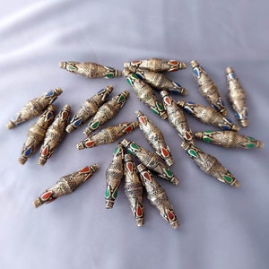 Puede incluir: Una colección de cuentas de metal dorado con diseños intrincados y acentos de esmalte de colores. Las cuentas están dispuestas en un patrón disperso sobre una superficie gris claro.