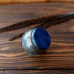 Peut inclure: Une bague en argent avec une pierre ovale en lapis-lazuli sertie au centre. La bague est ornée d'un motif gravé complexe autour de la pierre.