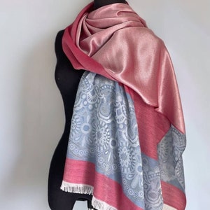 May include: A pink and gray patterned scarf with a fringe on the bottom edge. The scarf is made of a soft, silky material.
