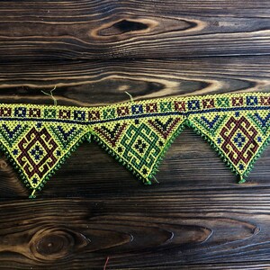 Puede incluir: Una guirnalda de cuentas con un patrón geométrico amarillo, verde, rojo y azul. La guirnalda está hecha de cuentas pequeñas y está enhebrada en forma triangular.
