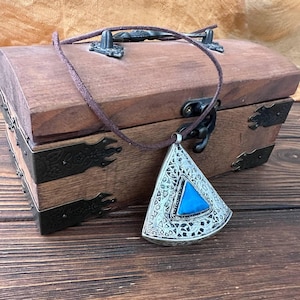 Puede incluir: Un colgante triangular de tono plateado con un intrincado patrón de filigrana y una piedra azul central. El colgante cuelga de un cordón marrón fino, descansando sobre un cofre de madera rectangular con bisagras y cierre de metal oscuro.