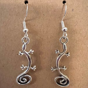 May include: A pair of silver dangle earrings featuring a stylized lizard design. The lizard is curled into a spiral shape.