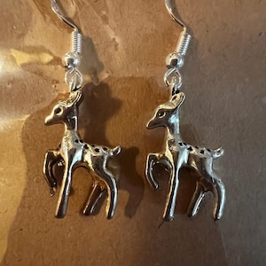 以下が含まれることがあります： 銀色の鹿のイヤリングのペア。各イヤリングは、銀色のフックから吊り下げられた、小さな詳細な鹿のペンダントが特徴です。鹿は、足、角、斑点が目立つようにデザインされています。