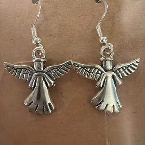 May include: A pair of silver-colored angel earrings. Each earring features a detailed angel figure with outstretched wings and a flowing gown. The earrings have a simple hook design for easy wear.