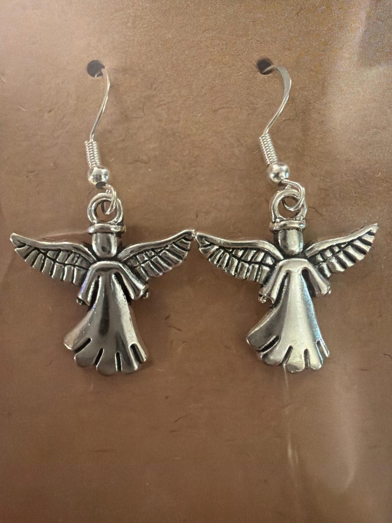 May include: A pair of silver-colored angel earrings. Each earring features a detailed angel figure with outstretched wings and a flowing gown. The earrings have a simple hook design for easy wear.