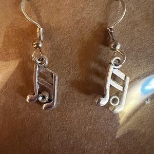 May include: A pair of silver-toned earrings featuring a musical eighth note design. Each earring has a curved hook, a small bead, and a spring coil detail. The musical note has a small, clear gem in the center.